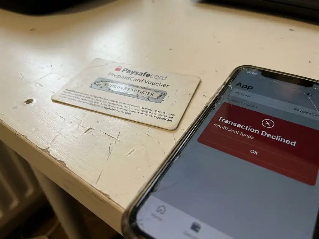 Abgekratzter PaysafeCard-Gutschein auf weißem Tisch neben Smartphone mit abgelehnter Transaktion.
